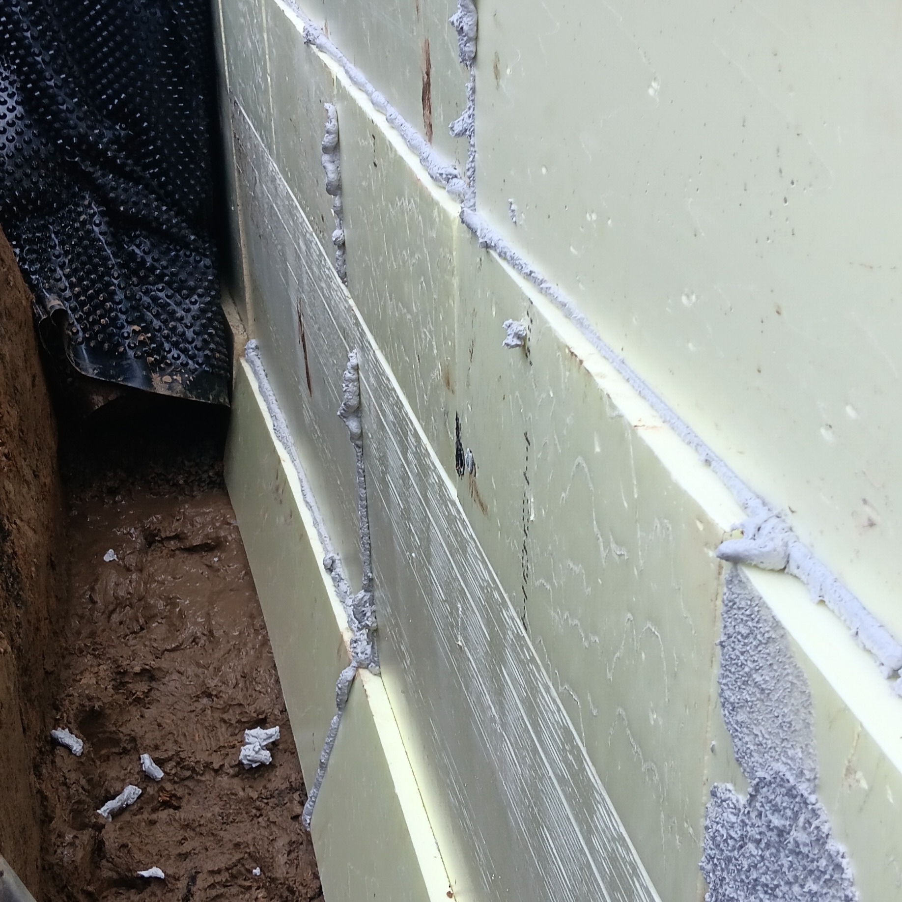 Ocieplenie fundamentu z użyciem styropianu, zabezpieczone folią kubełkową. Widoczne łączenia płyt styropianowych wypełnione pianką montażową. Grunt wokół fundamentu.