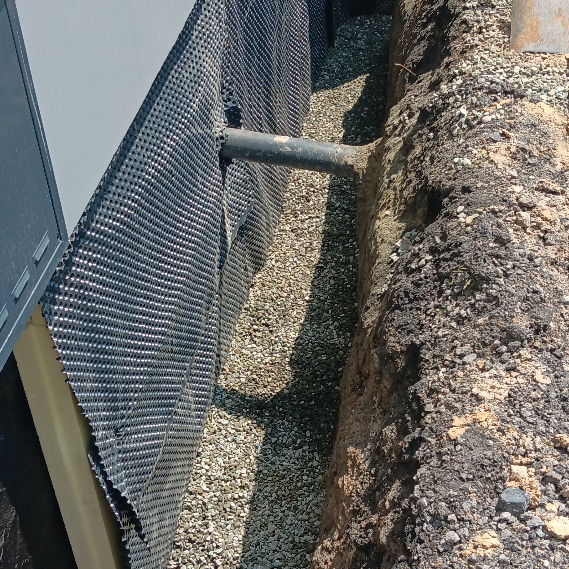 Izolacja pionowa fundamentu z folią kubełkową i drenażem żwirowym, rura odprowadzająca wodę. Przekrój wykopu, widoczne warstwy gruntu.