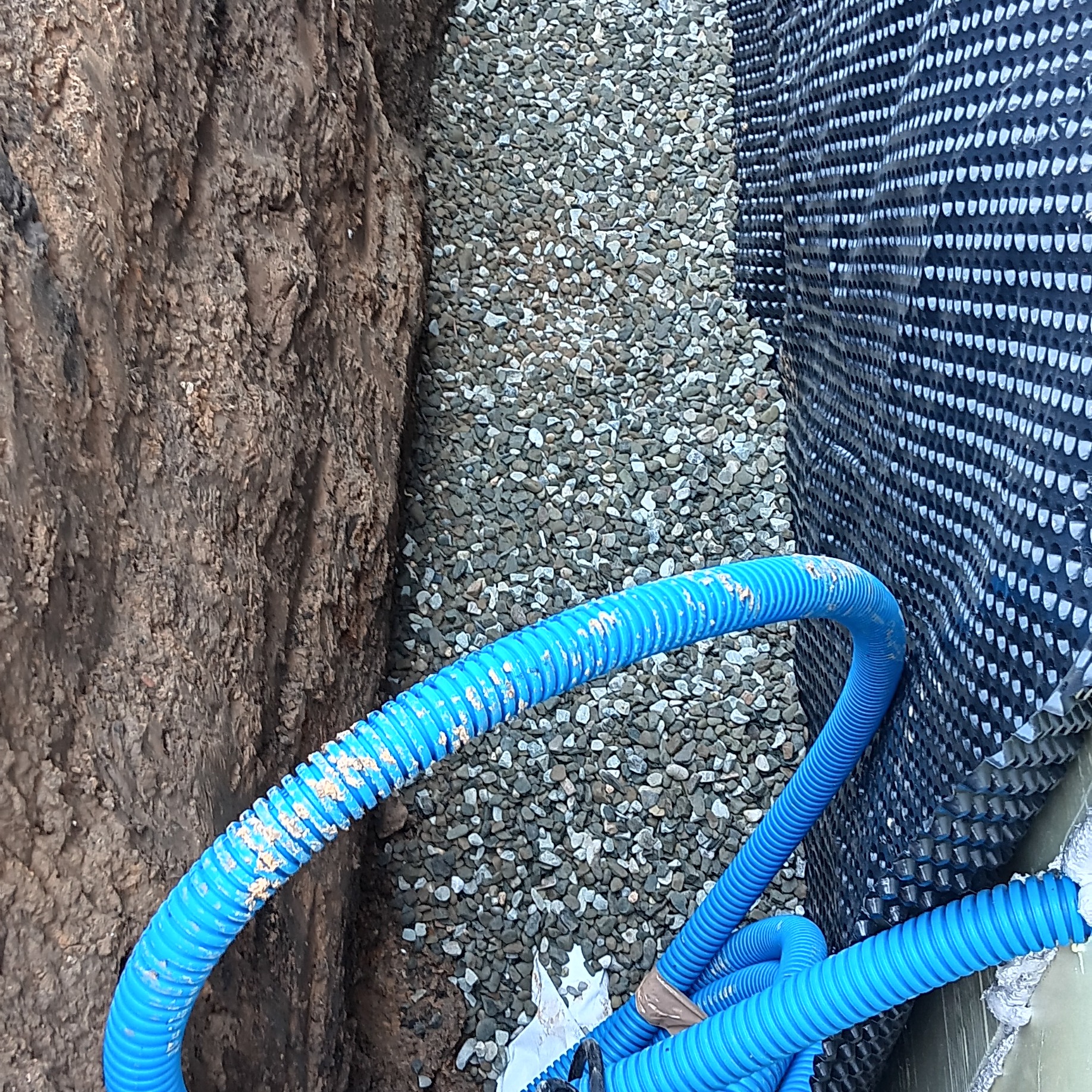 Wykop fundamentowy z drenażem: brązowa ziemia, szare kamienie, niebieskie rury drenażowe i czarna folia izolacyjna. Widok z góry na drenaż fundamentów.