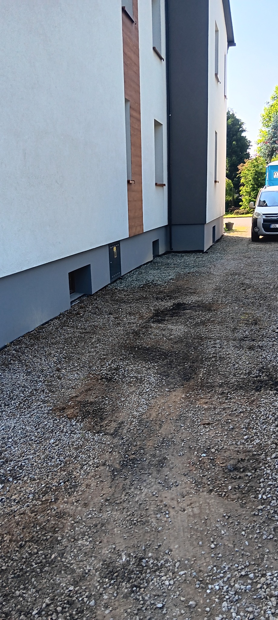 Budynek z nową elewacją i utwardzonym terenem wokół, widoczne okienka piwniczne i samochód w tle. Utwardzona droga żwirowa.