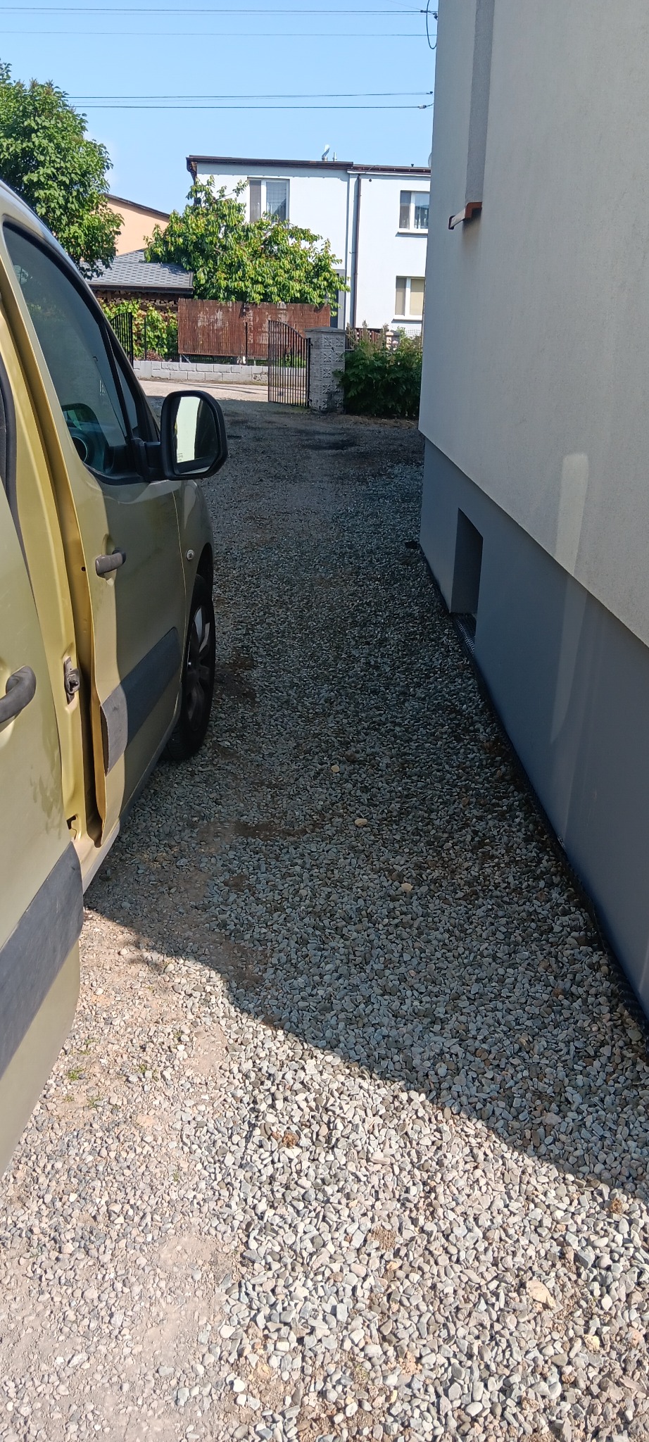 Żwirowa ścieżka obok budynku, widok z boku samochodu. Ścieżka prowadzi do bramy, w tle zabudowa jednorodzinna. Słoneczny dzień.