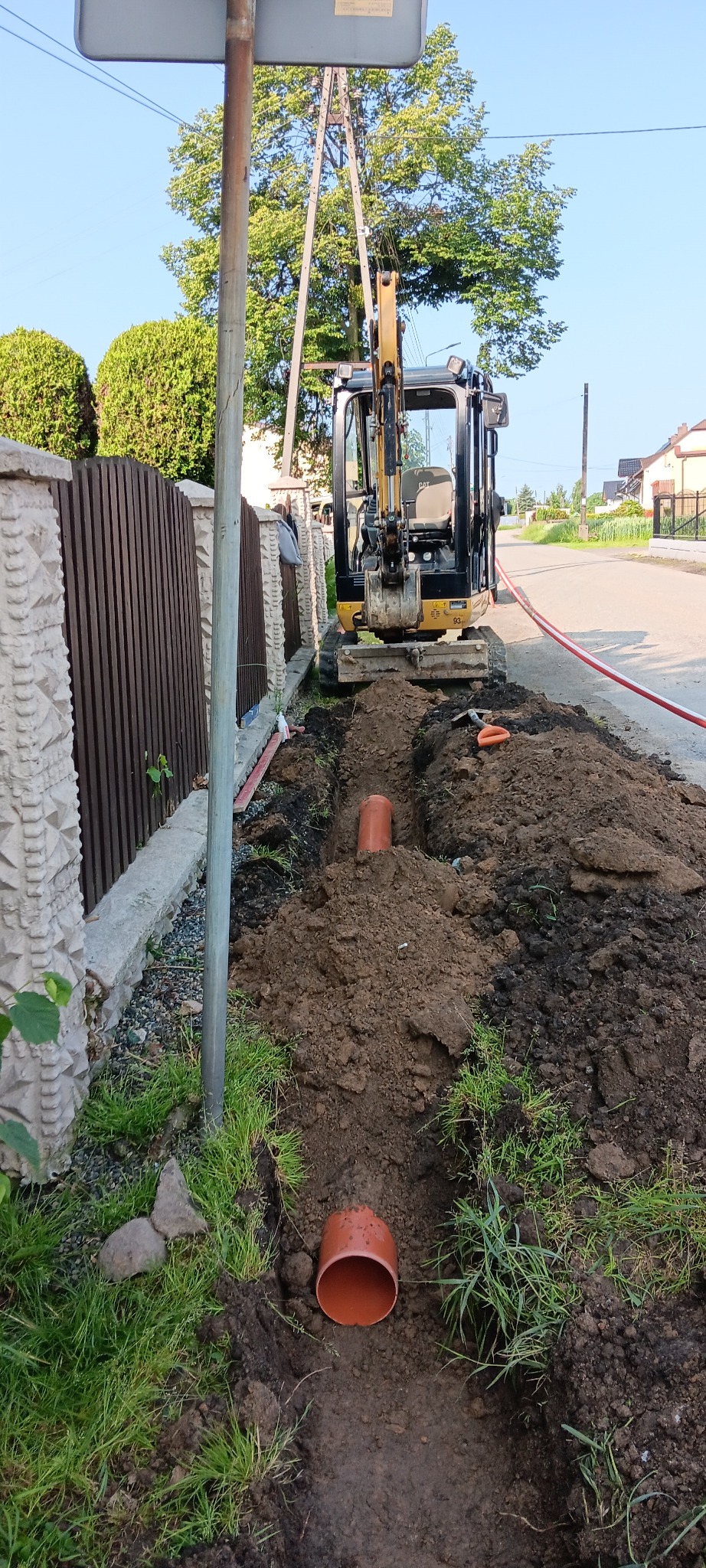 Mini koparka przy wykopie pod rury kanalizacyjne, widok z góry na rów z pomarańczowymi rurami, ogrodzenie z prawej, słup energetyczny z lewej.