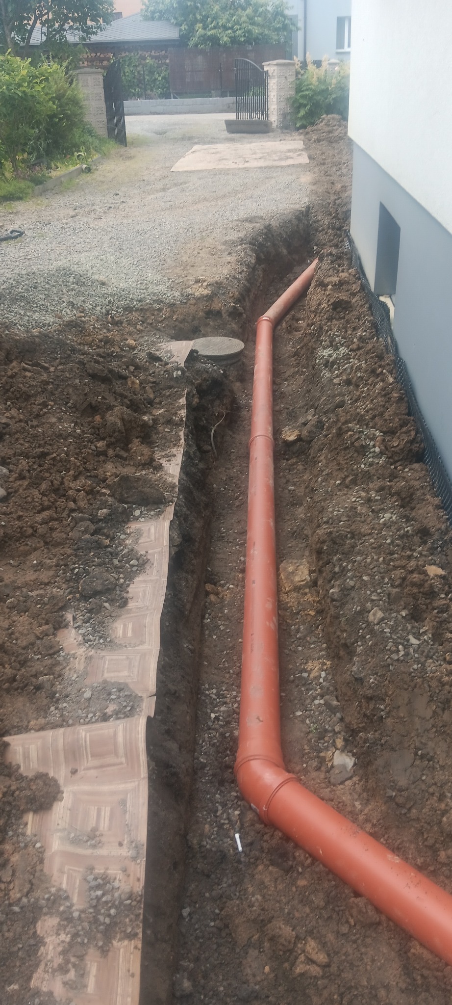Pomarańczowa rura drenażowa ułożona w wykopie wzdłuż fundamentu domu, zabezpieczona geowłókniną. Widoczny fragment posesji z bramą w tle.
