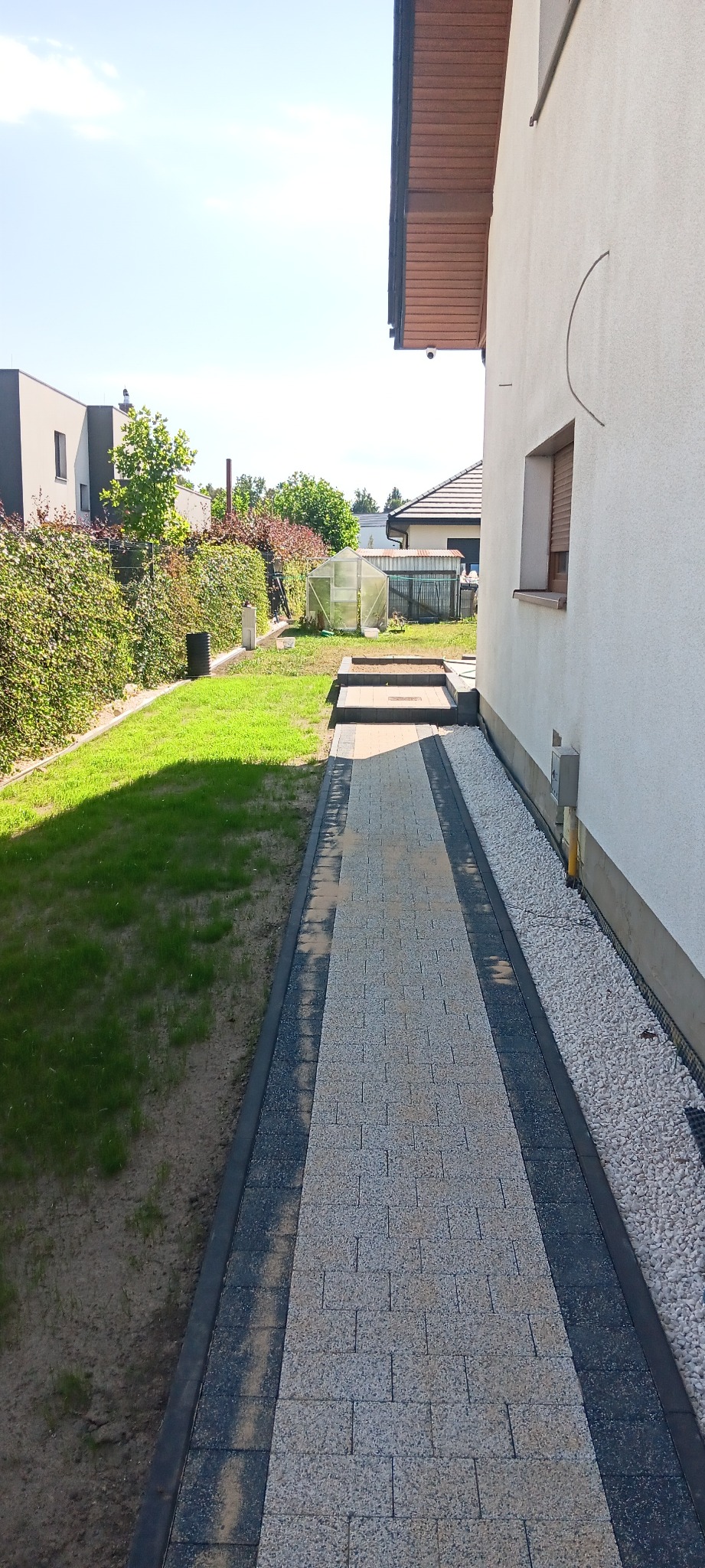 Ścieżka z kostki brukowej, obrzeża z ciemnej kostki, po prawej stronie biały żwir przy ścianie domu, po lewej trawnik i zielony żywopłot. Słoneczny dzień.