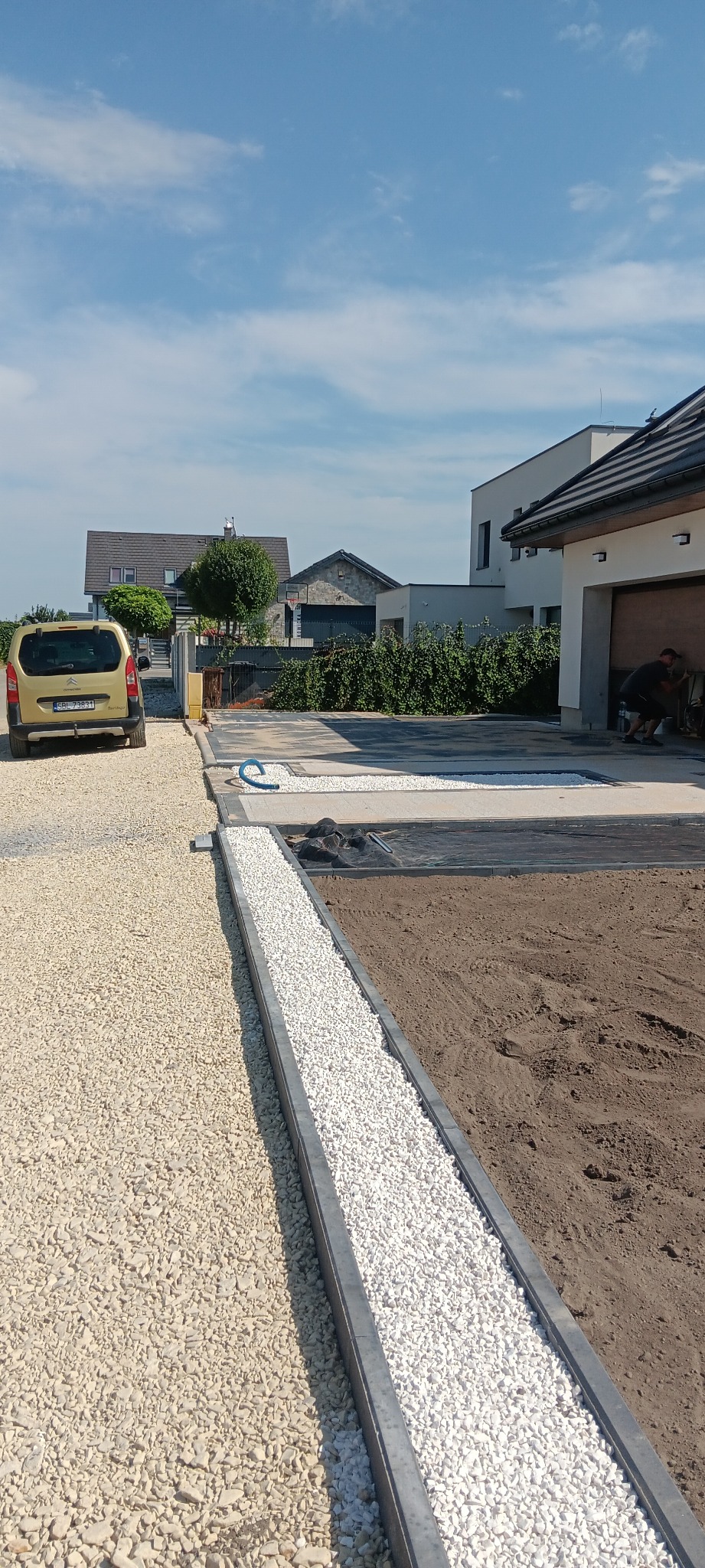 Wykończony podjazd z jasnego żwiru i białych kamyków oddzielonych szarym obrzeżem. W tle dom z garażem i zaparkowany samochód.