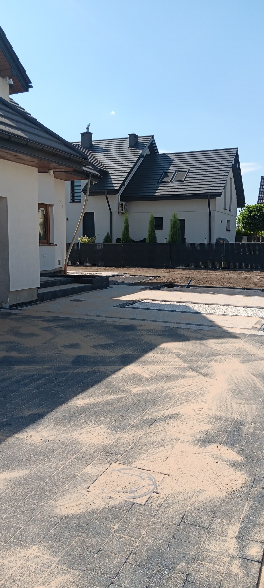 Nowoczesna posesja z ułożoną kostką brukową w odcieniach szarości, z widocznym włazem kanalizacyjnym. W tle domy z ciemnymi dachami i ogrodzeniem.