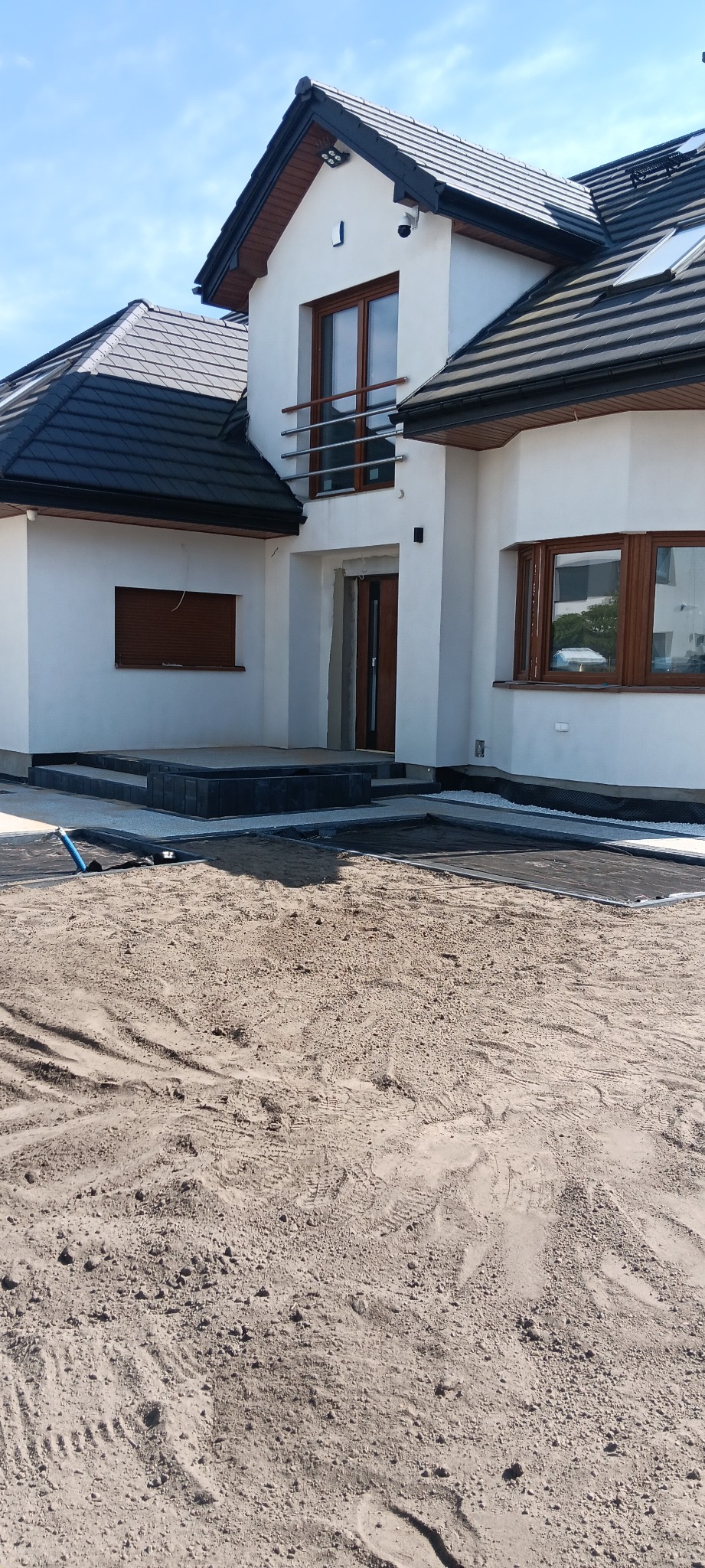 Nowoczesny dom jednorodzinny z wykończonym tarasem i przygotowanym terenem pod dalsze prace brukarskie. Widoczne czarne obrzeża i podsypka.