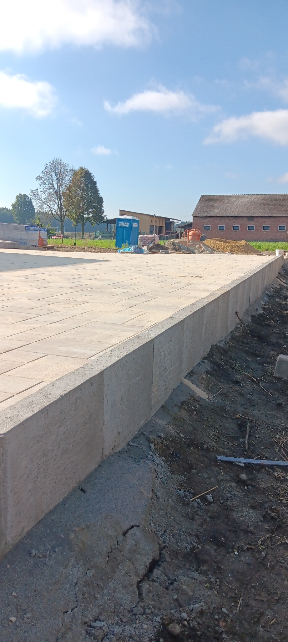 Ukończona nawierzchnia z kostki brukowej na przygotowanym podłożu. Widoczny mur oporowy, w tle budynek gospodarczy i błękitne niebo z chmurami.