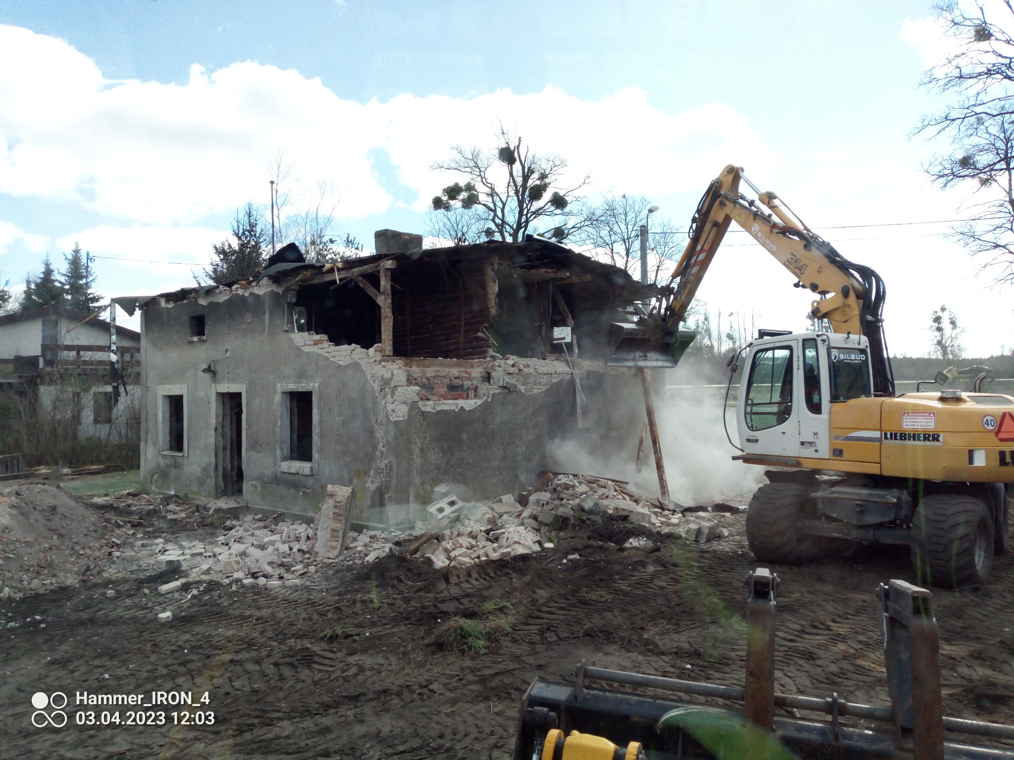 Żółta koparka Liebherr 314 z wysięgnikiem kruszy resztki szarego, częściowo rozebranego budynku z drewnianą konstrukcją dachu, w tle błękitne niebo z chmurami.