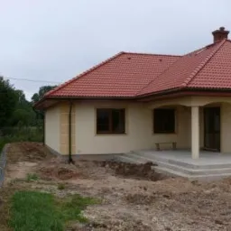 Jednopiętrowy dom z czerwoną dachówką, świeżo otynkowaną elewacją w odcieniach beżu i kremu, z widocznymi fragmentami ocieplenia na ścianach i przygotowanym terenem wokół budynku.