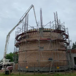 Wznoszenie okrągłej, murowanej konstrukcji z cegły, otoczonej rusztowaniami i zbrojeniami, z pompą do betonu w tle, na placu budowy.