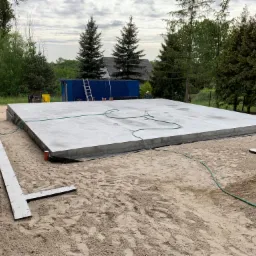 Wylana płyta fundamentowa na piasku z widocznym szalunkiem i izolacją, na tle drzew i kontenera budowlanego.