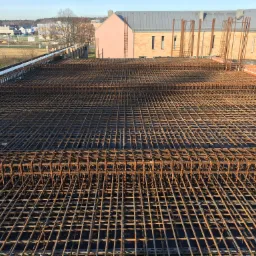 Siatka zbrojeniowa ułożona na deskowaniu, przygotowana do zalania betonem, widoczne pionowe pręty zbrojeniowe wystające ponad siatkę, w tle budynki mieszkalne.