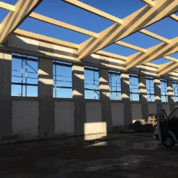 Wnętrze w trakcie budowy hali z elementami żelbetowymi i drewnianą konstrukcją dachu, widoczne ściany z bloczków, częściowo otynkowane, oraz dostawczy samochód z otwartymi drzwiami.