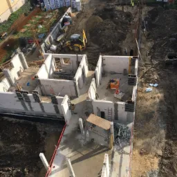 Budowa domu z betonu komórkowego widziana z góry, z wykopami, fundamentami, ścianami parteru, stertami materiałów budowlanych i pracującą koparką.