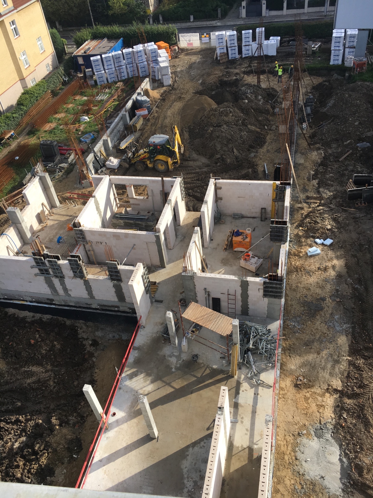 Budowa domu z betonu komórkowego widziana z góry, z wykopami, fundamentami, ścianami parteru, stertami materiałów budowlanych i pracującą koparką.