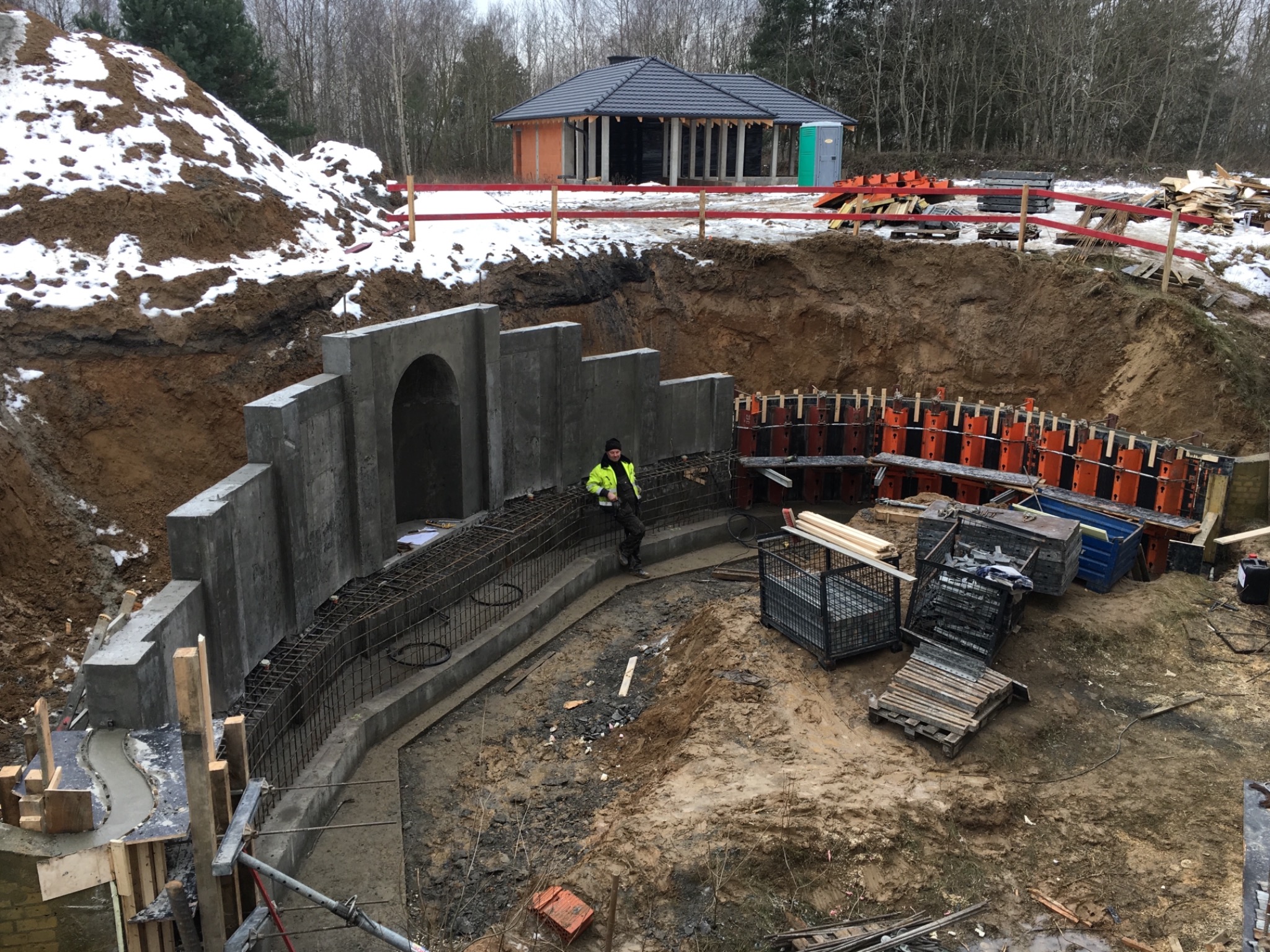 Budowa betonowej konstrukcji z łukiem w wykopie, widoczne zbrojenia, szalunki i pracownik w odblaskowej kurtce. W tle budynek z kolumnami i sterta ziemi z resztkami śniegu.