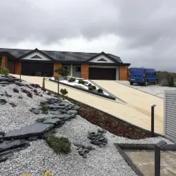 Nowoczesny dom z dwoma garażami, podjazd wyłożony jasną kostką, zagospodarowany teren wokół domu z kamieniami i roślinami, niebieski samochód dostawczy zaparkowany na podjeździe.