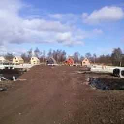 Fundamenty pod domy w zabudowie szeregowej, częściowo w trakcie budowy. W tle widoczne niebieskie niebo i drzewa. Żuraw budowlany w lewym górnym rogu.