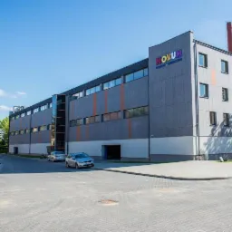 Nowoczesny budynek biurowy z szarą elewacją, pomarańczowymi akcentami i logo 'NOVUM' na fasadzie. Dwa samochody zaparkowane przed budynkiem w słoneczny dzień.