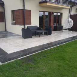 Wyłożony jasnymi płytami taras z ciemną obwódką, na którym stoi czarny komplet mebli ogrodowych i białe wiszące krzesło, przylegający do beżowego domu z brązowymi okiennicami.