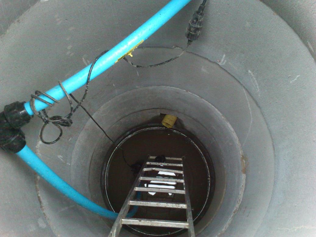 Wnętrze betonowej studni z wodą, widoczna metalowa drabina, niebieskie rury z podłączeniem elektrycznym i element systemu drenarskiego.