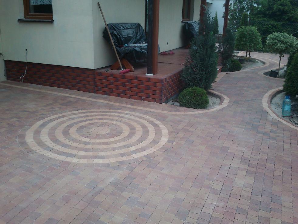 Wykończony podjazd z kostki brukowej w odcieniach czerwieni i beżu, z dekoracyjnym wzorem okręgów, prowadzący do tarasu i ścieżki w ogrodzie.