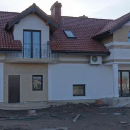 Dwukondygnacyjny dom jednorodzinny w stanie surowym zamkniętym, z czerwoną dachówką, elewacją z białego tynku i fragmentami wykończenia imitującego kamień, widoczne okno okrągłe i balkon z czarną...