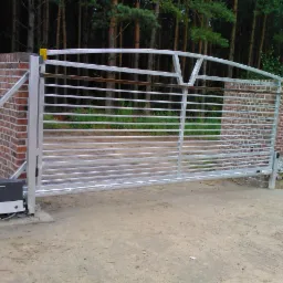 Nowoczesna, przesuwna brama posesyjna ze stali ocynkowanej, z automatyką, na tle ceglanego muru i lasu. Widoczny napęd bramy.