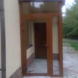 Zewnętrzne drzwi wejściowe z aluminiową ramą w kolorze brązowym, widoczne z zewnątrz budynku na tle kostki brukowej.