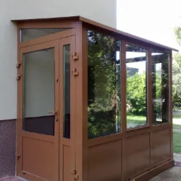 Brązowa, aluminiowa zabudowa wejścia z przeszklonymi ścianami i drzwiami, widok z zewnątrz, ceglana ścieżka prowadzi do wejścia.