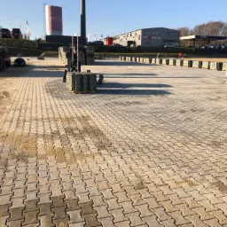 Ułożona kostka brukowa w kształcie zygzaka na dużym placu, z żółtą koparką i paletami kostki w tle, w pobliżu stacji benzynowej i budynku Moto Strefa.