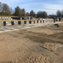 Przygotowanie terenu pod budowę drogi z paletami kostki brukowej w tle, widoczny walec i pracownicy.