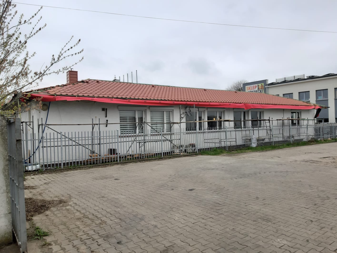 Budynek z czerwonym dachem w trakcie naprawy, zabezpieczony folią i otoczony rusztowaniem.