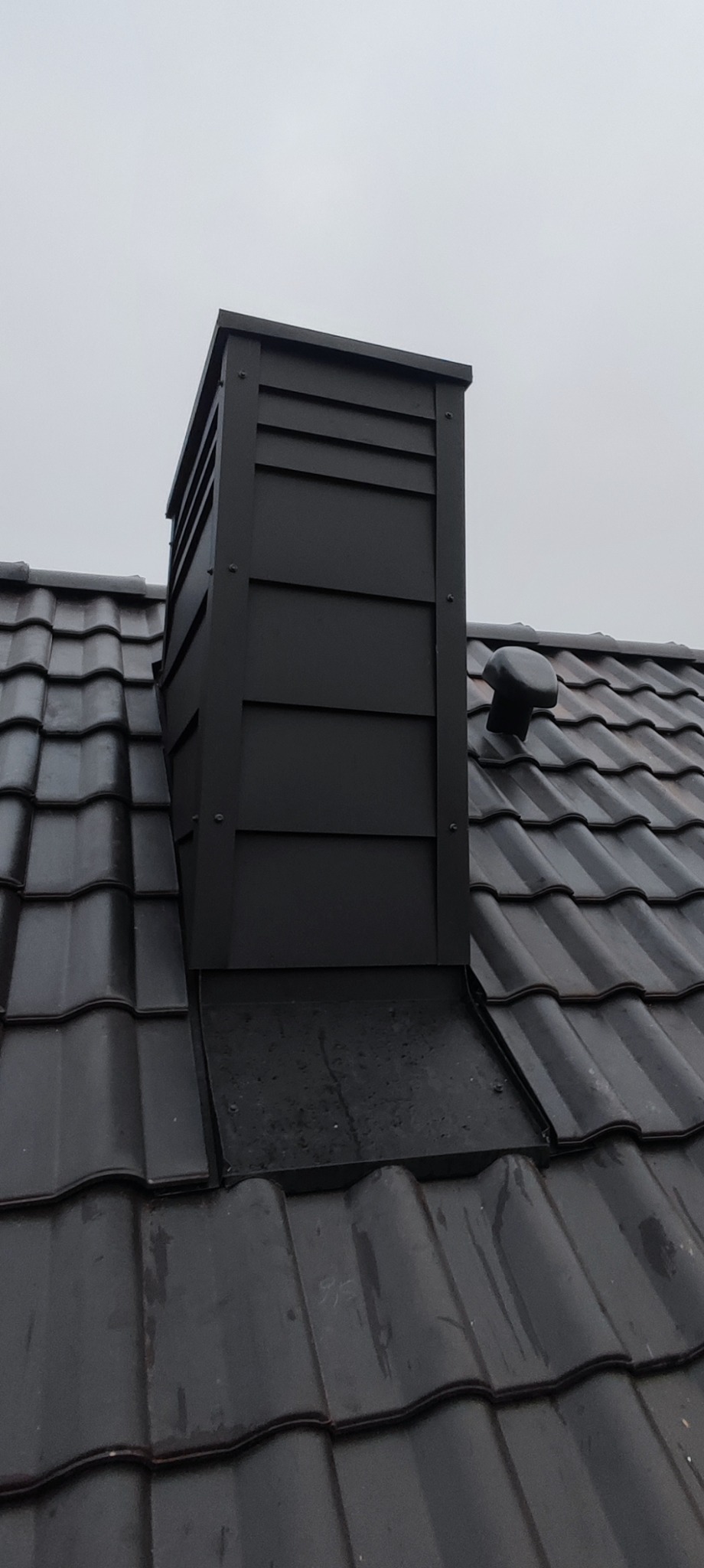Nowoczesny, czarny komin wentylacyjny z metalowymi panelami na błyszczącym, ciemnoszarym dachu ceramicznym w pochmurny dzień.