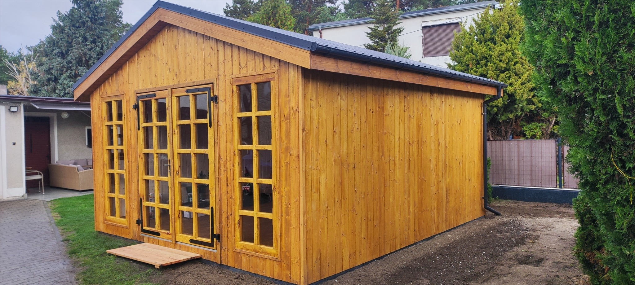 Drewniana altana z przeszklonymi drzwiami i czarnym dachem, usytuowana na trawniku. Widoczne drzewa w tle i fragment domu. Naturalne oświetlenie.