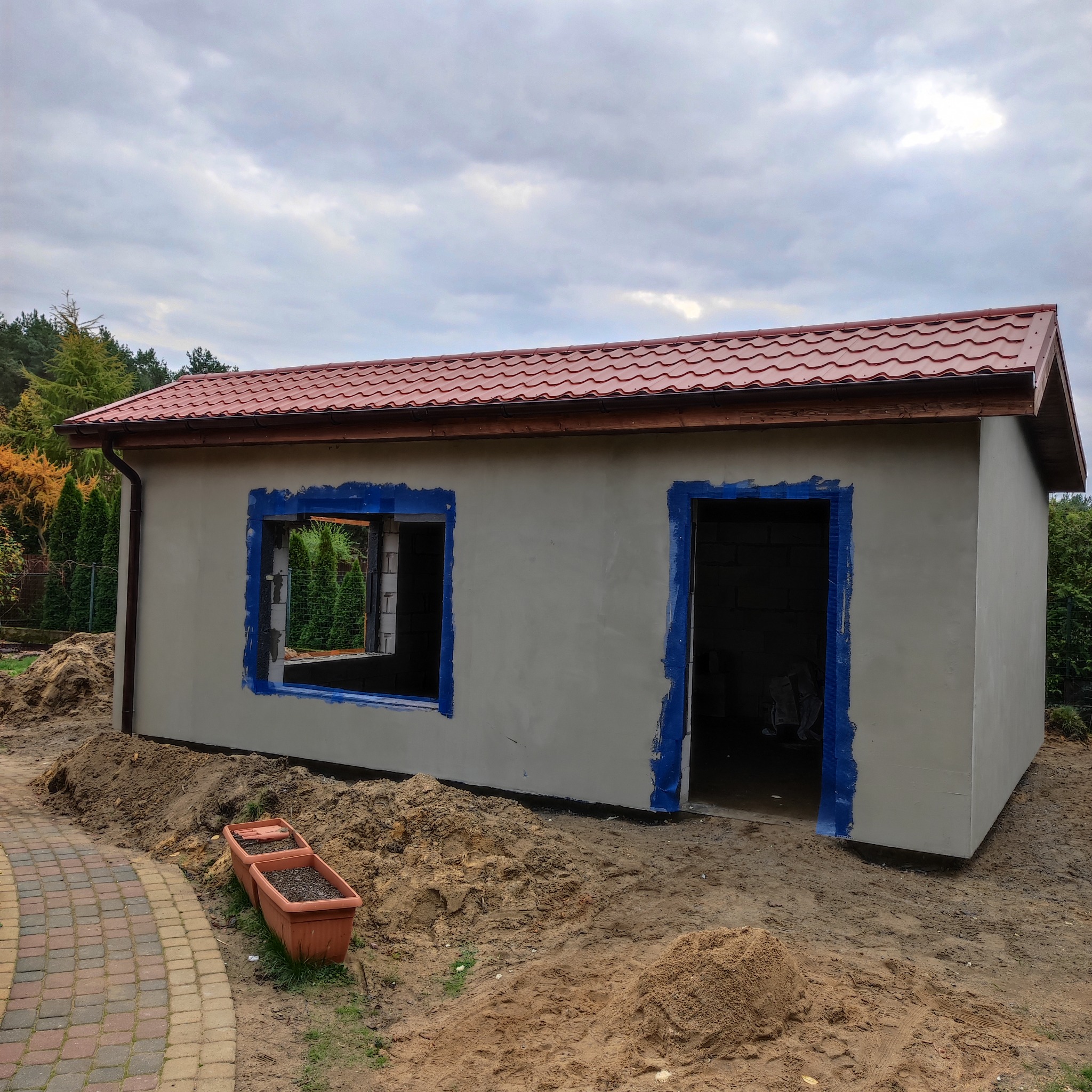 Niedokończony budynek gospodarczy z czerwoną blachodachówką, zabezpieczonymi na niebiesko otworami okiennymi i drzwiowymi, otoczony ziemią i roślinnością.