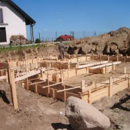 Przygotowanie szalunków pod fundamenty domu jednorodzinnego z widocznym zbrojeniem, na tle ziemi i domu z drewnem opałowym.