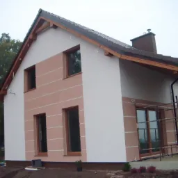 Dwukondygnacyjny dom z jasną elewacją w odcieniach beżu i bieli, z drewnianą konstrukcją werandy i rusztowaniem przy oknie, na tle pochmurnego nieba.