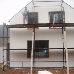 Dwukondygnacyjny dom w trakcie prac elewacyjnych: robotnik na rusztowaniu z płytą izolacyjną, okna zabezpieczone folią, widoczny fragment ocieplonej ściany budynku gospodarczego.