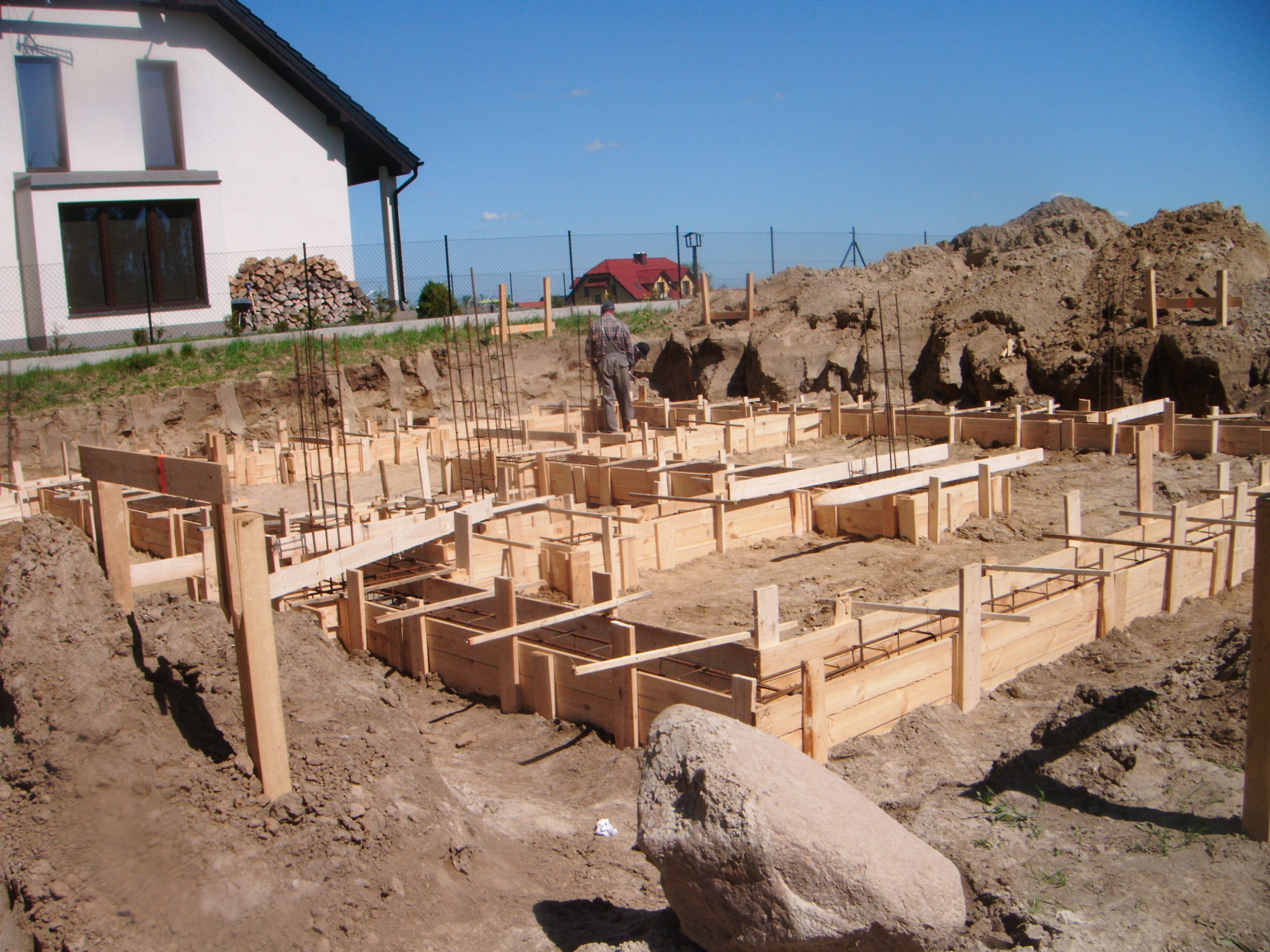 Przygotowanie szalunków pod fundamenty domu jednorodzinnego z widocznym zbrojeniem, na tle ziemi i domu z drewnem opałowym.