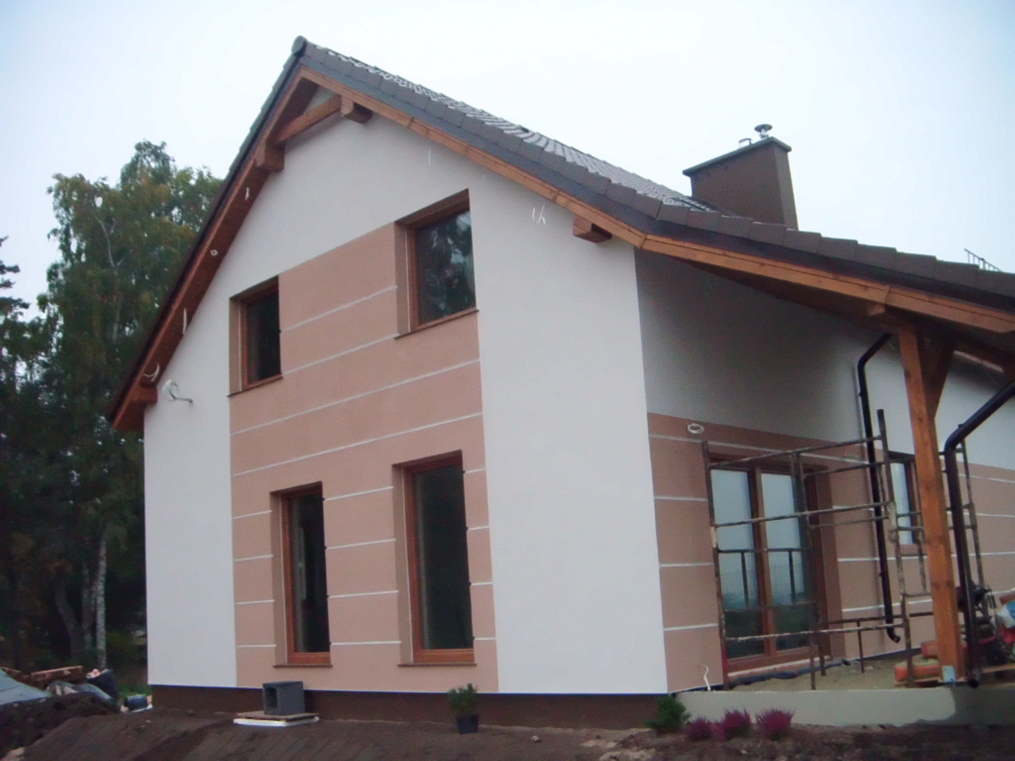 Dwukondygnacyjny dom z jasną elewacją w odcieniach beżu i bieli, z drewnianą konstrukcją werandy i rusztowaniem przy oknie, na tle pochmurnego nieba.