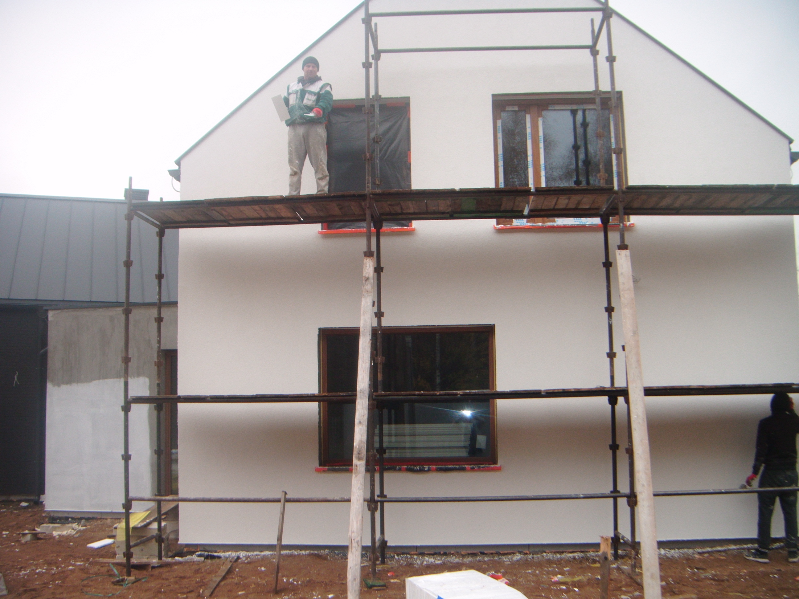 Dwukondygnacyjny dom w trakcie prac elewacyjnych: robotnik na rusztowaniu z płytą izolacyjną, okna zabezpieczone folią, widoczny fragment ocieplonej ściany budynku gospodarczego.