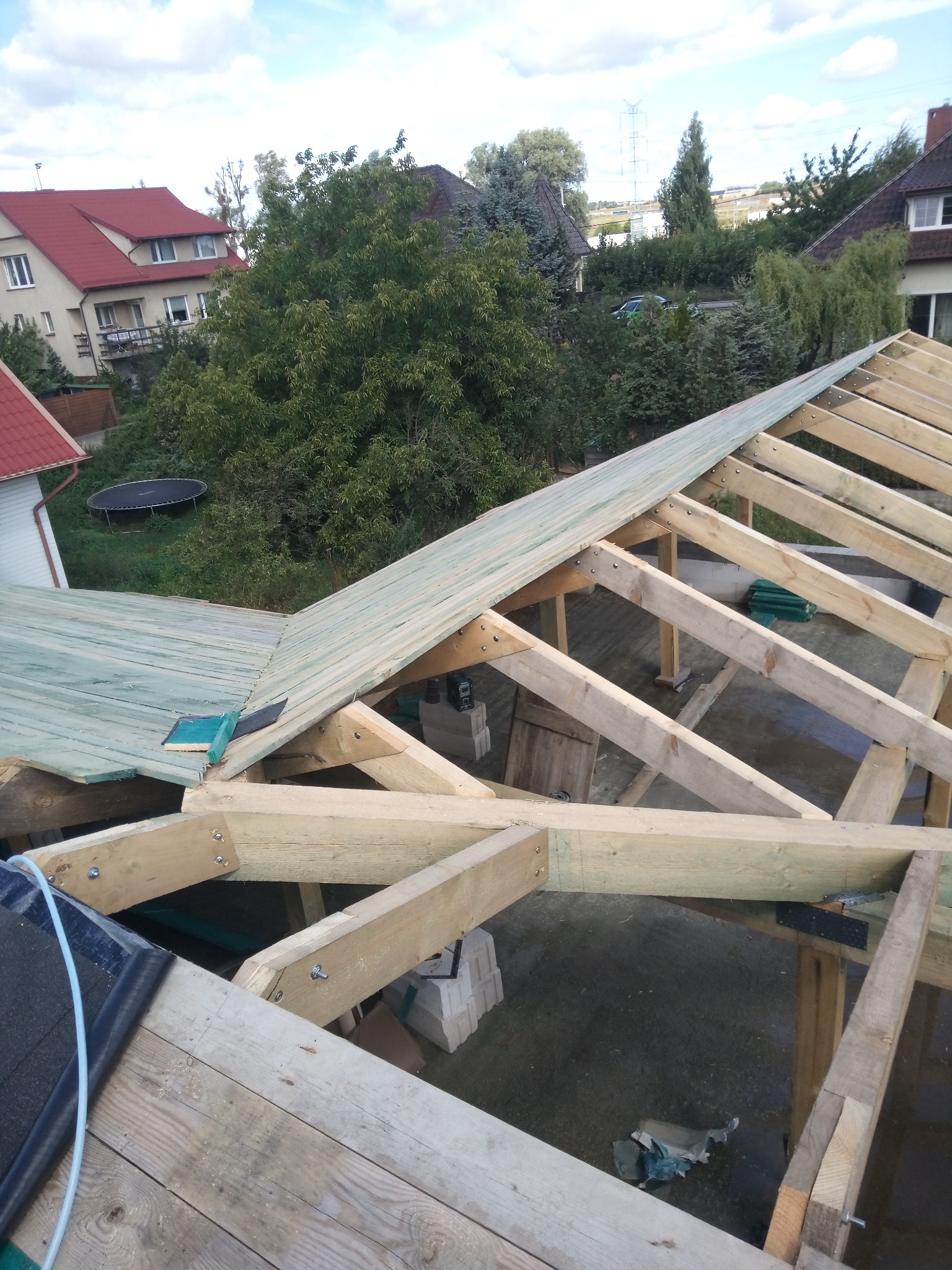 Wznoszenie drewnianej więźby dachowej z deskowaniem, widok z góry na konstrukcję z jasnego drewna na tle zieleni i zabudowań. Częściowo pokryte papą.