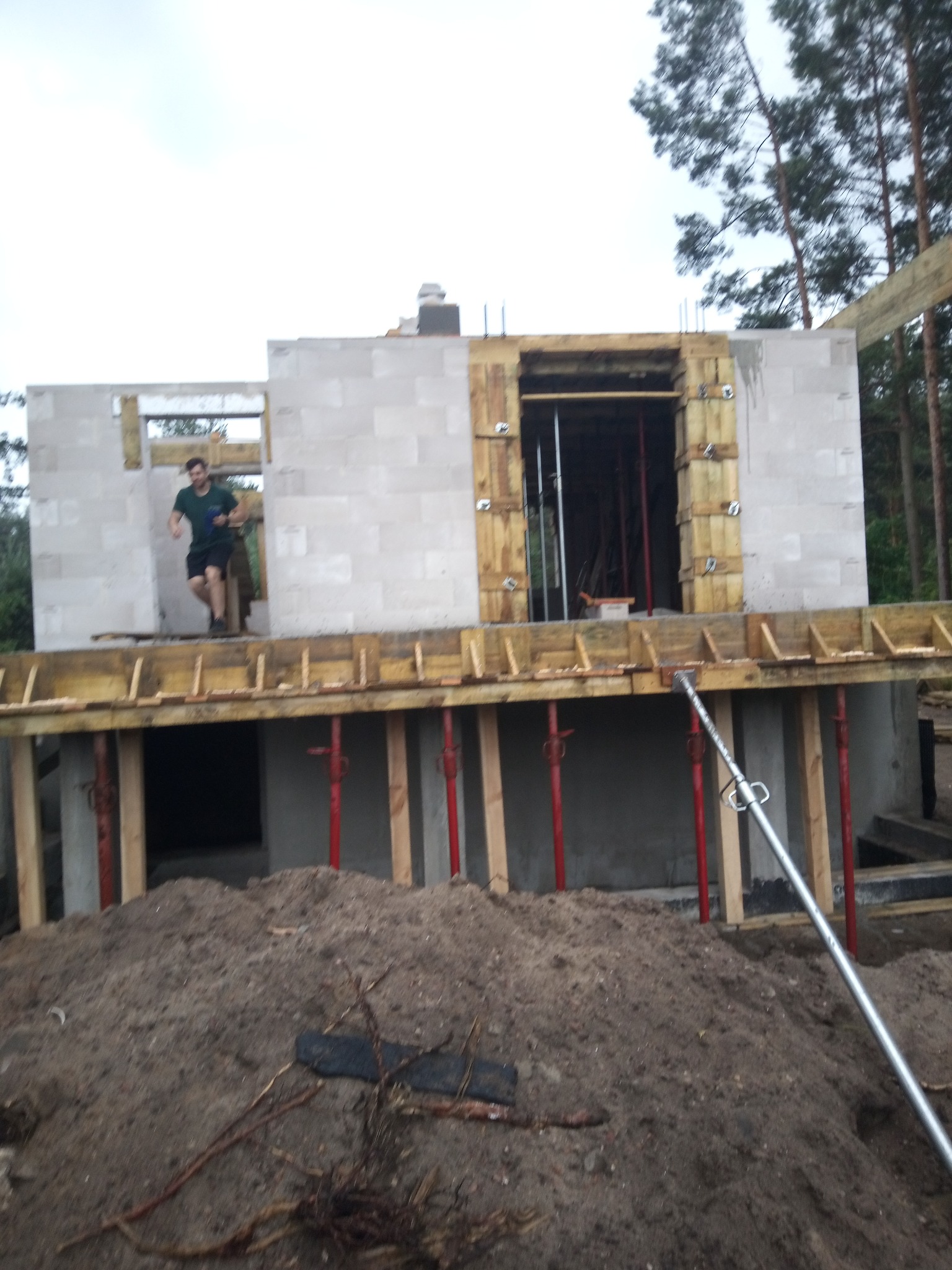 Wznoszenie ścian z bloczków betonu komórkowego na fundamencie. Widoczny pracownik w otworze okiennym, drewniane szalunki i podpory. W tle drzewa.