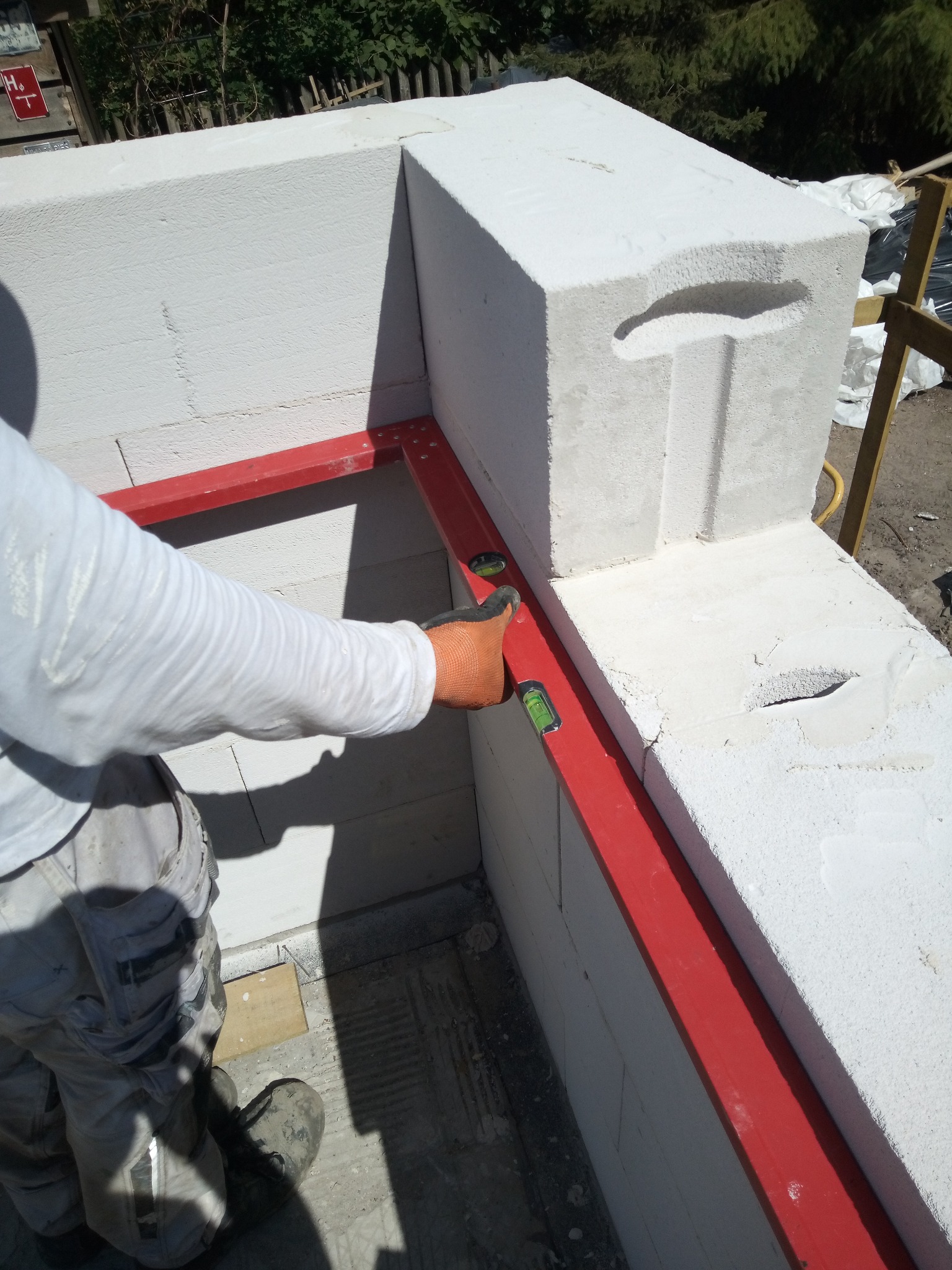 Murowanie ściany z białych bloczków. Murarz z poziomicą w dłoni sprawdza poziom. Czerwona listwa poziomująca. Widoczne detale struktury bloczków.