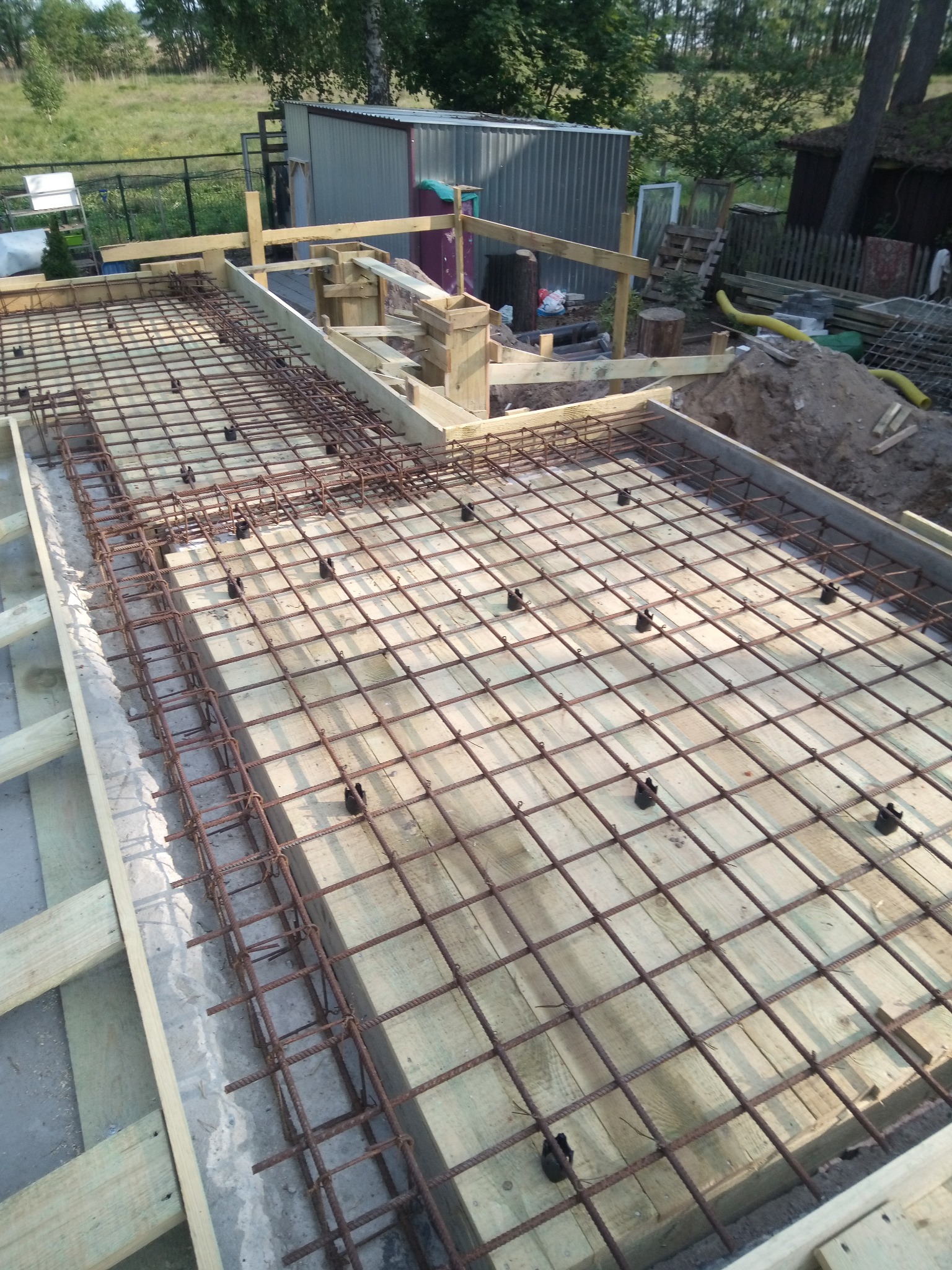 Zbrojenie fundamentu na deskowaniu z drewnianych desek, widoczne czarne dystanse. W tle ziemia, budynek gospodarczy i drzewa. Ujęcie z góry.