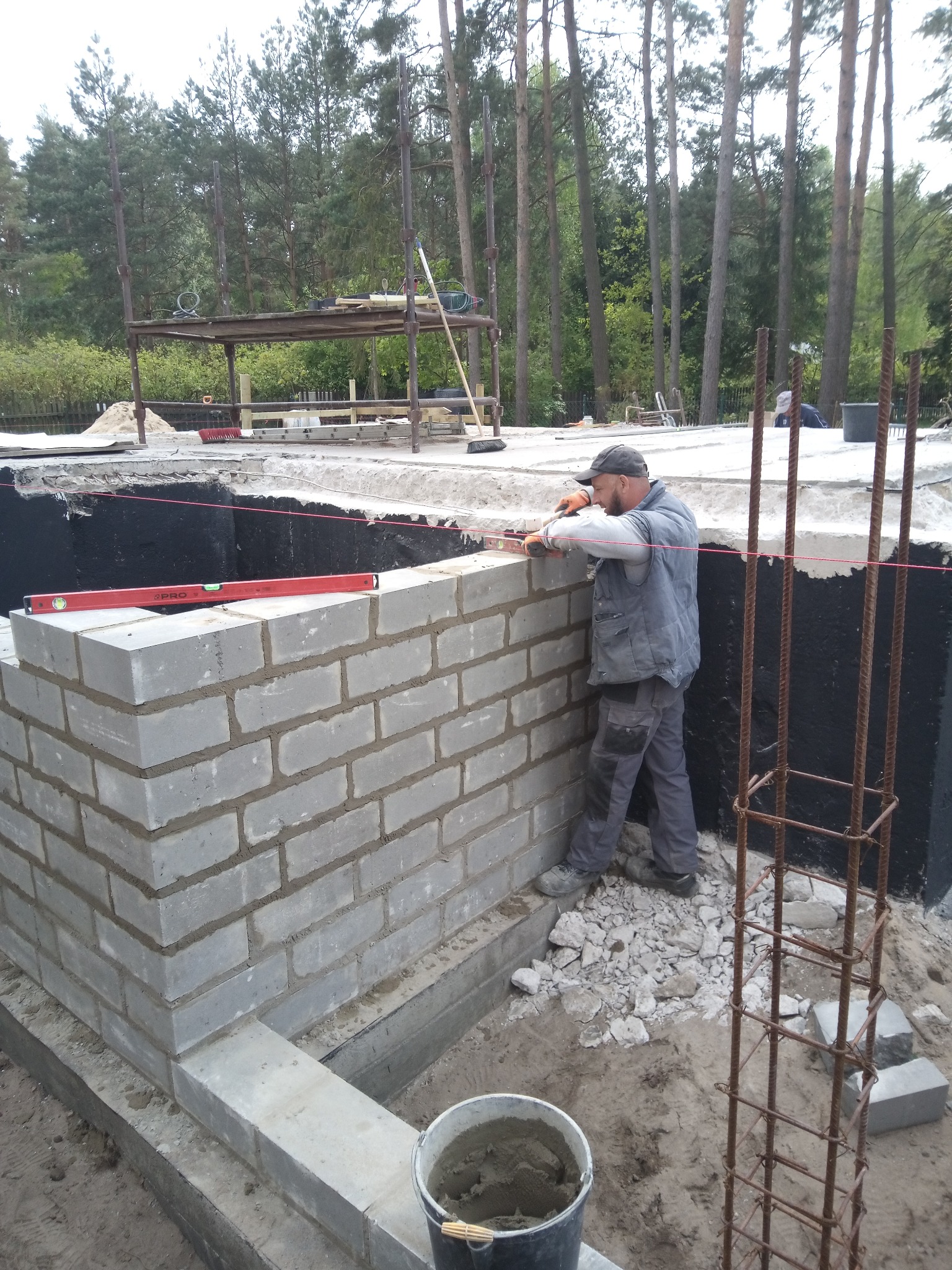 Murarz z poziomicą układa bloczki fundamentowe na fundamencie z izolacją. Widoczne zbrojenie, wiadro z zaprawą i las w tle.