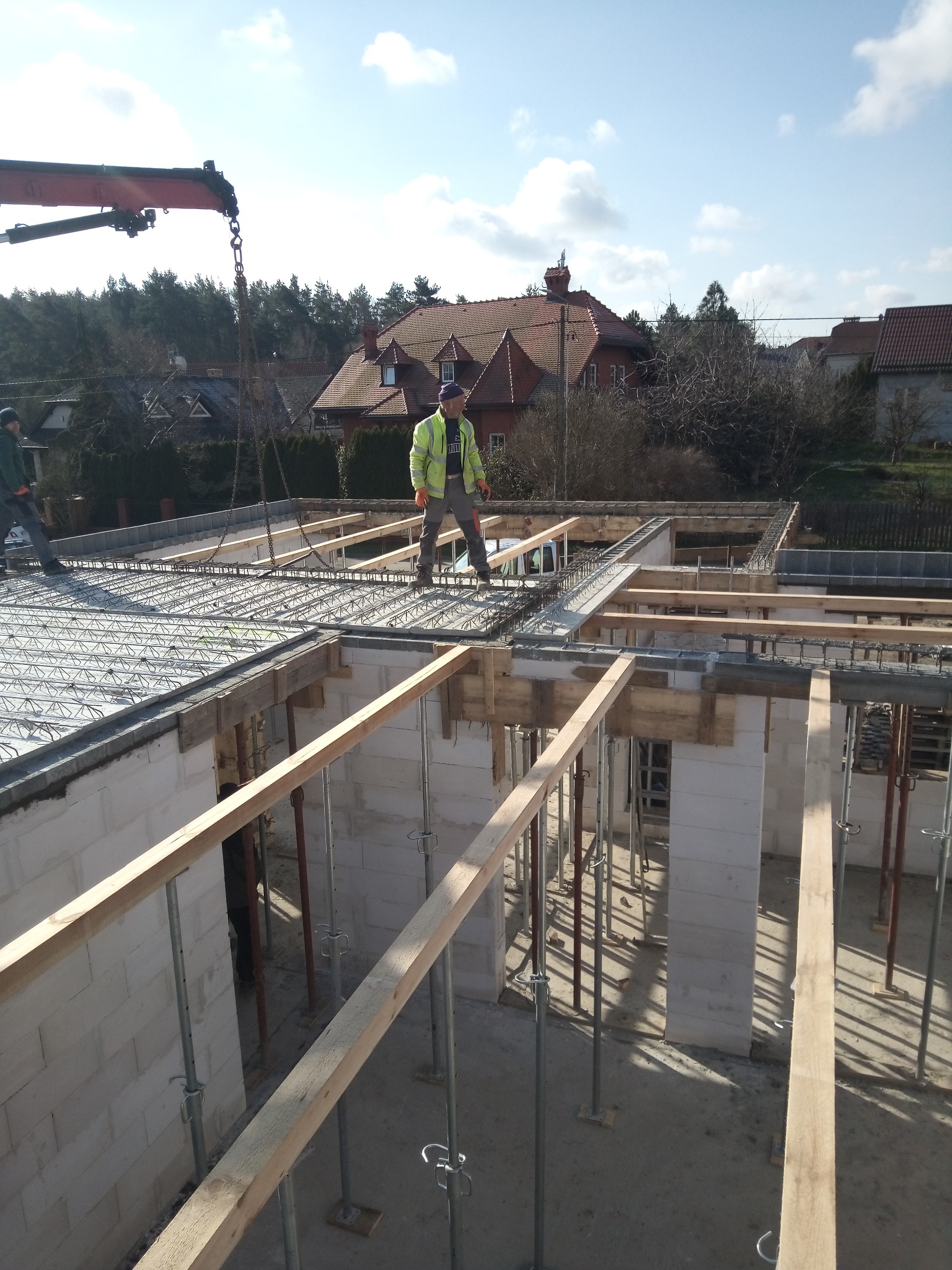 Budowa domu: robotnik w żółtej kurtce stoi na zbrojeniu stropu, widoczne białe bloczki fundamentowe, drewniane podpory i żuraw budowlany w tle.