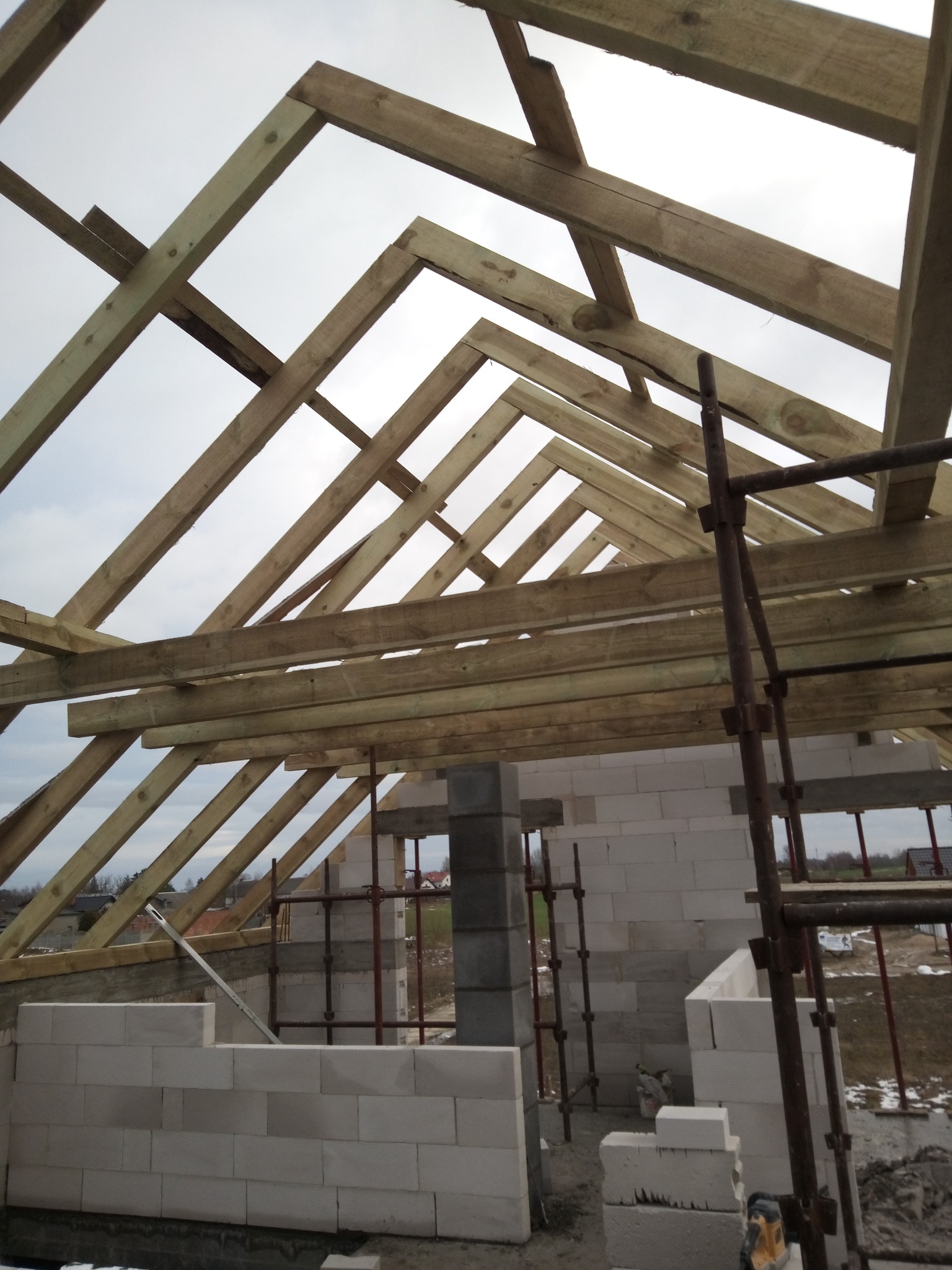 Wznoszenie drewnianej więźby dachowej na murowanych ścianach z bloczków, widok z dołu. Fragment rusztowania. W tle pochmurne niebo i zabudowania.
